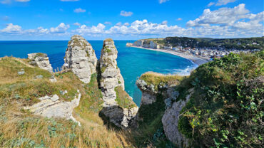 Atemberaubender Blick von einer hohen Klippe auf die Steilküste von Étretat in der Normandie. Im Vordergrund ragen markante Kalksteinfelsen mit grasbewachsenen Plateaus senkrecht aus dem türkisblauen Meer. Im Hintergrund erstreckt sich eine geschwungene Bucht mit feinem Kiesstrand und dem charmanten Küstenort Étretat. Der Himmel ist leuchtend blau mit dekorativen weißen Wolken – perfektes Wetter für eine Wanderung entlang der Alabasterküste.