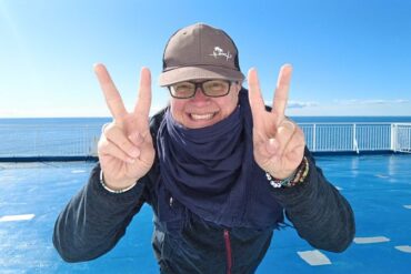 Frau mit Schal Jacke und Brille steht auf einem Schiffdeck und beugt sich zur Kamera. Sie strahlt übers ganze Gesicht und hält beide Hände mit Peacezeichen vor sich.