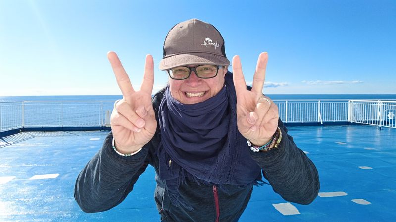 Frau mit Schal Jacke und Brille steht auf einem Schiffdeck und beugt sich zur Kamera. Sie strahlt übers ganze Gesicht und hält beide Hände mit Peacezeichen vor sich.