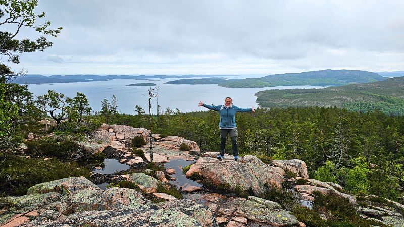 Frau steht mit ausgebreiteten Armen auf einem Felsplateau mit Blick auf das Meer und grüne Hügel in Schweden – Wandern in beeindruckender Naturkulisse.