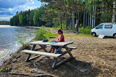 „Frau sitzt allein an einem Picknicktisch direkt am Seeufer in Schweden, im Hintergrund ein weißer Campervan am Waldrand – Szene voller Ruhe und Vanlife-Idylle.“