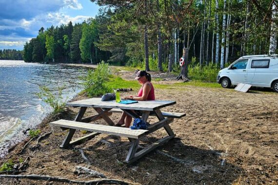 „Frau sitzt allein an einem Picknicktisch direkt am Seeufer in Schweden, im Hintergrund ein weißer Campervan am Waldrand – Szene voller Ruhe und Vanlife-Idylle.“