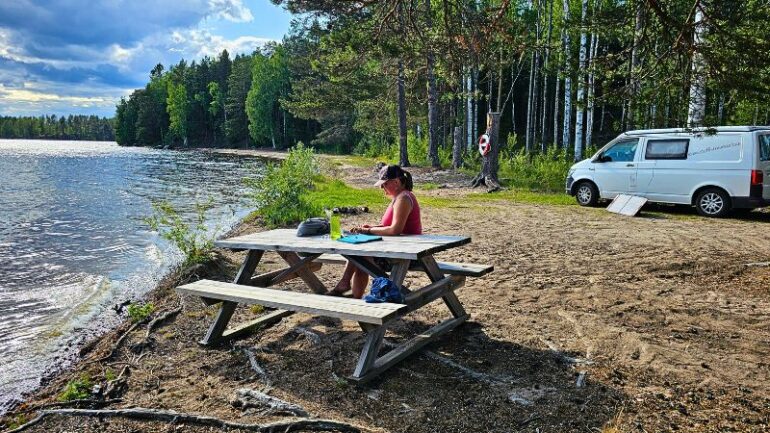 „Frau sitzt allein an einem Picknicktisch direkt am Seeufer in Schweden, im Hintergrund ein weißer Campervan am Waldrand – Szene voller Ruhe und Vanlife-Idylle.“