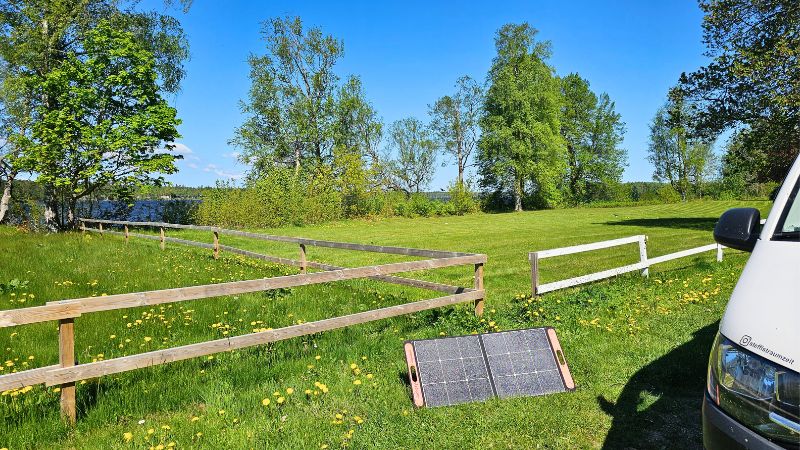 Eine grüne Wiese, Bäume, ein Zaun und ein Solarpanel das auf der Wiese Stegt. Auserdem sieht man ein Stückchen VW t& Campervan