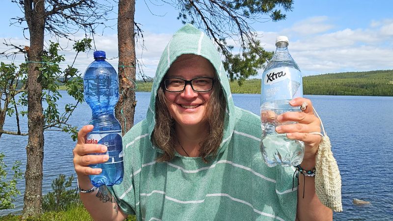 Frau mit Kapuzenponcho und Brille steht lachend vor einem See in Schweden, hält zwei große Wasserflaschen in den Händen – Vanlife-Alltag mit Humor.