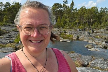 eine Frau mit Brille steht in einem pinken Trägertop in der Natur und lächelt in die Kamera