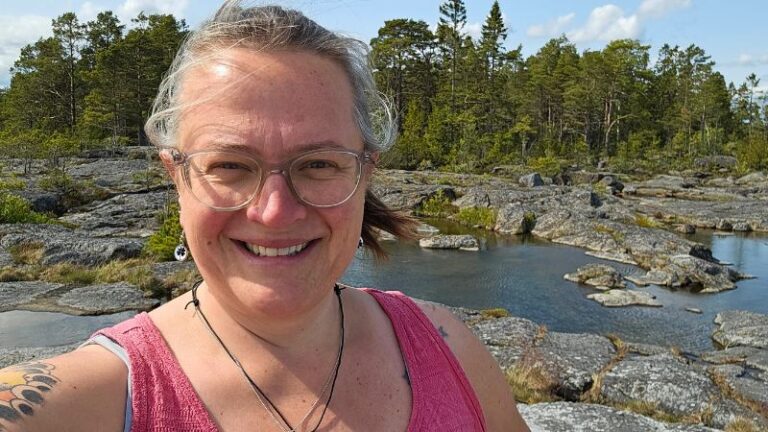 eine Frau mit Brille steht in einem pinken Trägertop in der Natur und lächelt in die Kamera