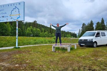 Frau steht auf einer Bank am Polarkreis. Auf der linken Seite sieht man das Polarkreisbild und rechts steht ein weißer VW T6