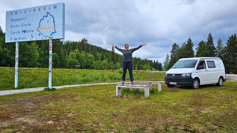 Frau steht auf einer Bank am Polarkreis. Auf der linken Seite sieht man das Polarkreisbild und rechts steht ein weißer VW T6