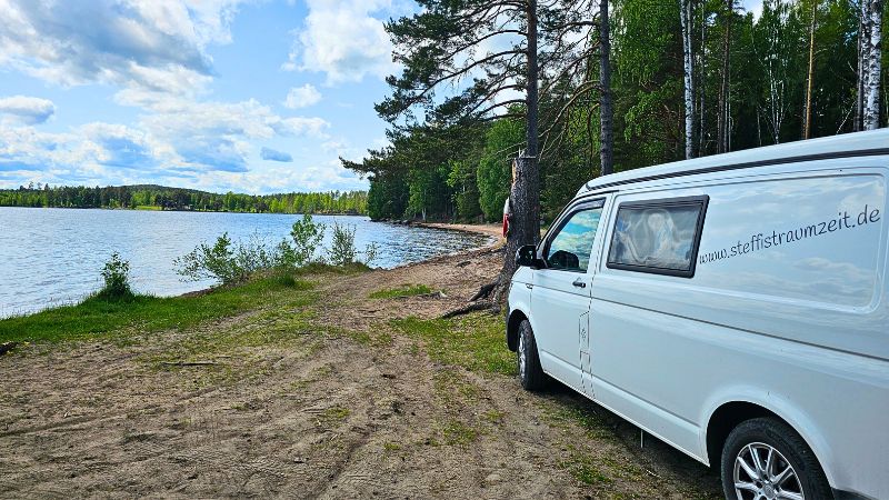 ein weißer VW T& steht an einem See. Man sieht einen kleinen Sanstrand und Wald