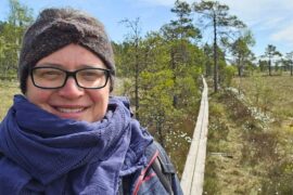 Frau mit Stirnband, Brille und dunkelblauem Schal lächelt in die Kamera – im Hintergrund verläuft ein Holzbohlenweg durch das offene Moor im Store Mosse Nationalpark, gesäumt von Wollgras und Kiefern.