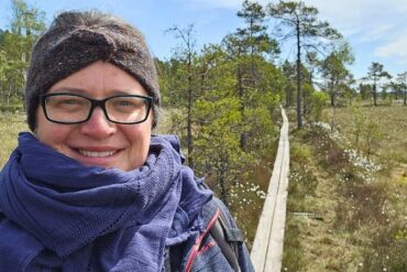 Frau mit Stirnband, Brille und dunkelblauem Schal lächelt in die Kamera – im Hintergrund verläuft ein Holzbohlenweg durch das offene Moor im Store Mosse Nationalpark, gesäumt von Wollgras und Kiefern.
