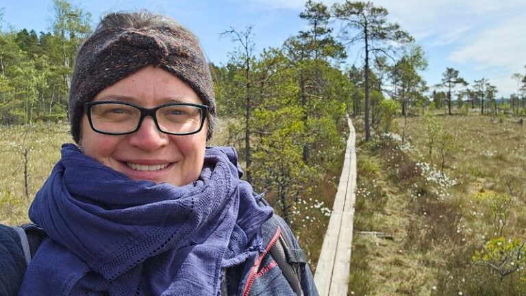 Frau mit Stirnband, Brille und dunkelblauem Schal lächelt in die Kamera – im Hintergrund verläuft ein Holzbohlenweg durch das offene Moor im Store Mosse Nationalpark, gesäumt von Wollgras und Kiefern.