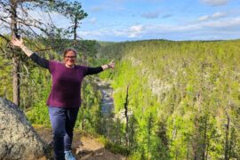 Frau mit ausgebreiteten Armen steht lachend auf einem Felsplateau mit Blick in eine tiefe, bewaldete Schlucht – im Hintergrund fließt ein Fluss durch das grüne Tal unter blauem Himmel mit Wolken.