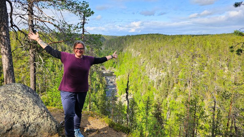 Frau mit ausgebreiteten Armen steht lachend auf einem Felsplateau mit Blick in eine tiefe, bewaldete Schlucht – im Hintergrund fließt ein Fluss durch das grüne Tal unter blauem Himmel mit Wolken.