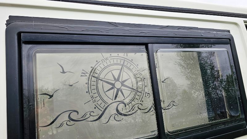Nahaufnahme eines Camperfensters mit dekorativer Folie: Windrose, Möwen und Wellenmotiv – oben ist schwarzes Dichtband zu sehen, das provisorisch ein undichtes Fenster abdichtet.