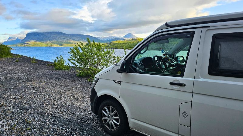 Weißer VW T6 Camper steht auf einem Schotterplatz direkt am Wasser – im Hintergrund beeindruckende Berglandschaft mit grünen Hügeln und schneebedeckten Gipfeln unter wolkigem Himmel.