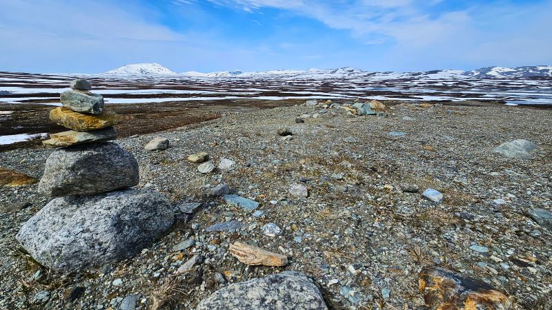 Steinmännchen aus flachen Felsen steht auf karger, steiniger Fläche – im Hintergrund schneebedeckte Berge und weite, offene Landschaft unter blauem Himmel mit Schleierwolken.