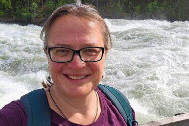 Frau mit Brille, violettem Shirt und Rucksack lächelt in die Kamera – im Hintergrund tosender Fluss mit starken Stromschnellen, umgeben von dichtem Wald.