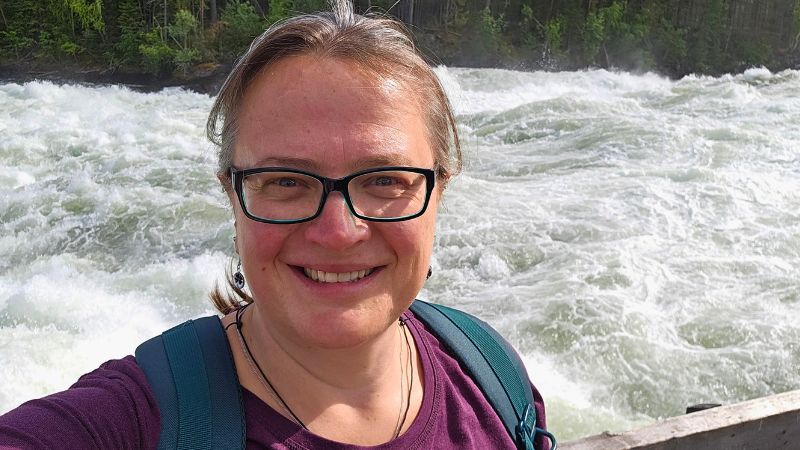 Frau mit Brille, violettem Shirt und Rucksack lächelt in die Kamera – im Hintergrund tosender Fluss mit starken Stromschnellen, umgeben von dichtem Wald.