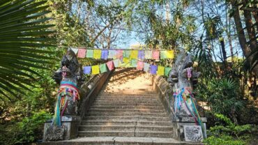 Eine Treppe in der Natur die hinaufführt und von bunten Gebetsfahnen überspannst ist. Auf jeder Treppenseite wacht ein Naga