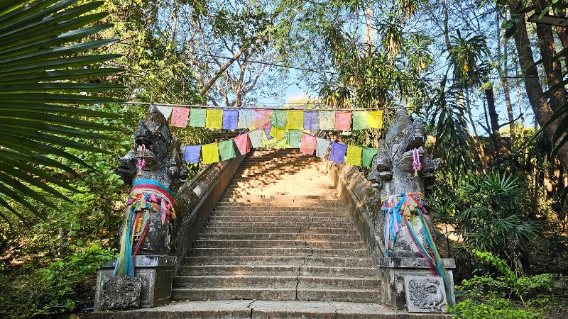 Eine Treppe in der Natur die hinaufführt und von bunten Gebetsfahnen überspannst ist. Auf jeder Treppenseite wacht ein Naga