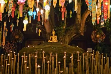 Ein golden Buddha auf einem Hügel unter einem Baum. Davor viele Kerzen auf Stäben. Am Baum hängen bunte Lampions