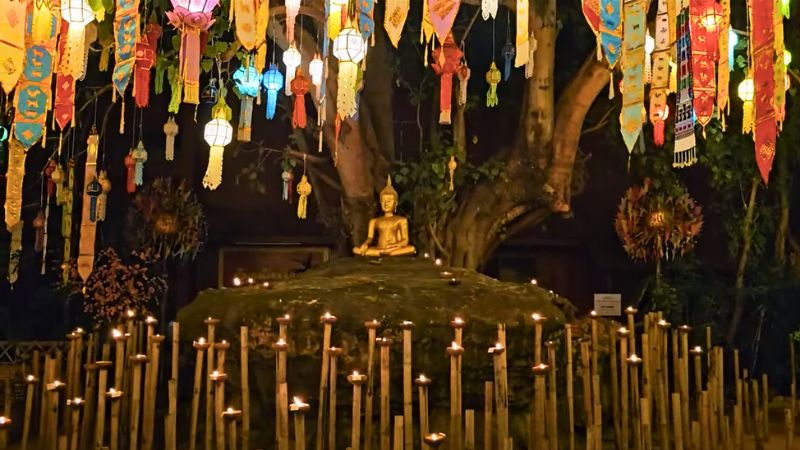 Ein golden Buddha auf einem Hügel unter einem Baum. Davor viele Kerzen auf Stäben. Am Baum hängen bunte Lampions