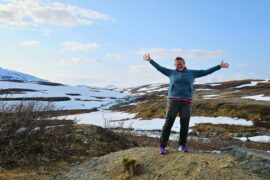 eine Frau steht mit ausgebreiteten armen in der Natur. im Hintergrund sieht man Hügel und teilweise noch Schnee