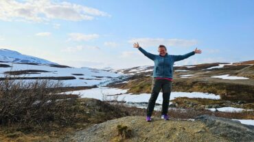 eine Frau steht mit ausgebreiteten armen in der Natur. im Hintergrund sieht man Hügel und teilweise noch Schnee