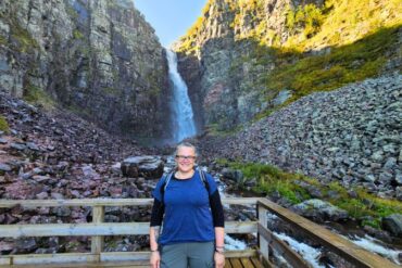 Eine Frau mit Brille steht lächelnd vor einem Wasserfall im Fulufjället Nationalpark