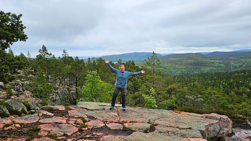 eine Frau steht mit ausgebreiteten Armen auf einem felsen und schaut lächeln in die Kamera. Im Hintergrund ist Wald zu sehen