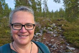 Eine Frau mit Brille lächelt in die Kamera. Im Hintergrund sieht man Bäume und einen Wanderweg