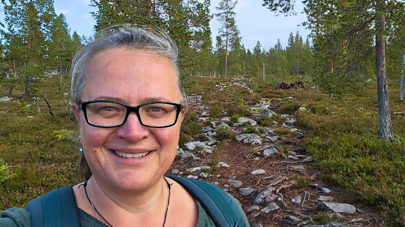 Eine Frau mit Brille lächelt in die Kamera. Im Hintergrund sieht man Bäume und einen Wanderweg